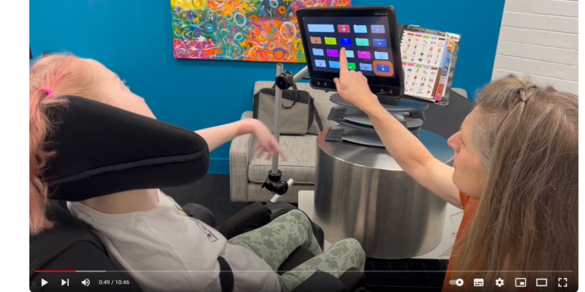 image description: viewing from behind over the shoulders of two people - person on left sitting in supportive wheelcahir with headrest and person on right looking at other person while pointing at screen of an electronic communication device - in coversation.