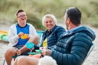 Down Syndrome young person on left sitting in camp chair, smiling and talking with parents to the right of image who also sit in amping chairs - outdoors