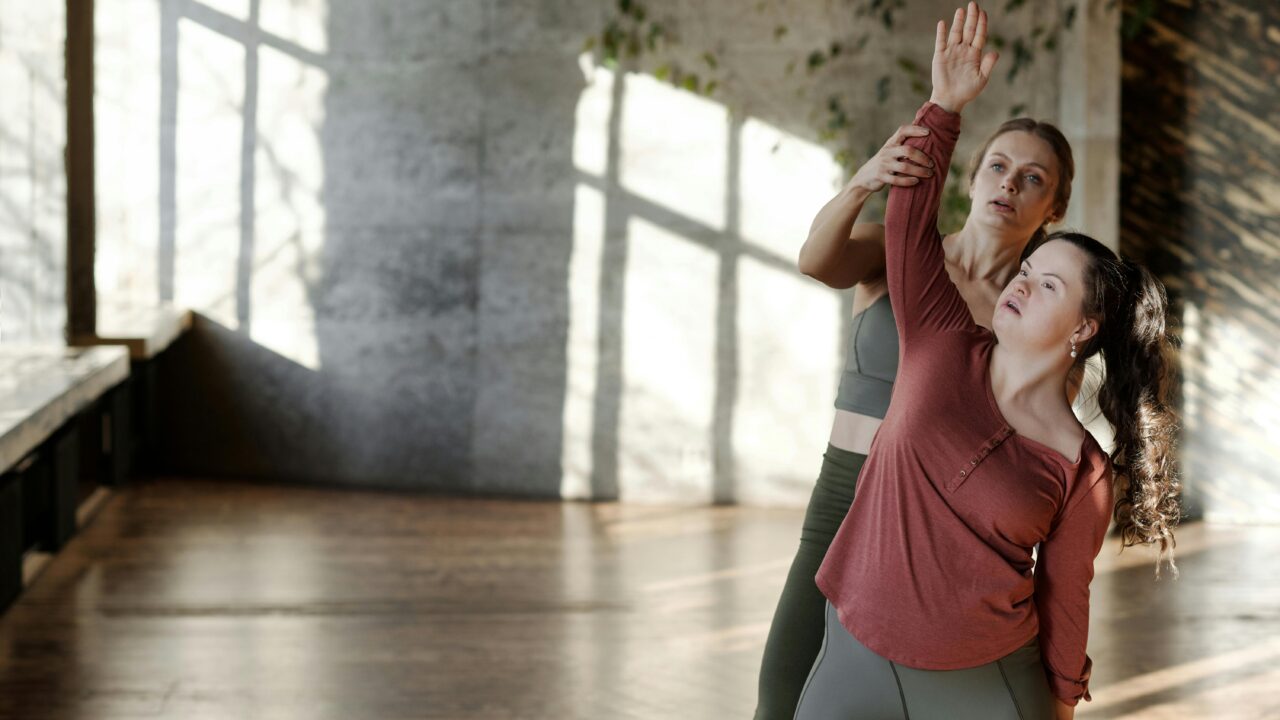 SUPPORT WORKER HELPING PERSON WITH DOWN SYNDROME IN A DANCE STUDIO WITH MOVEMENTS