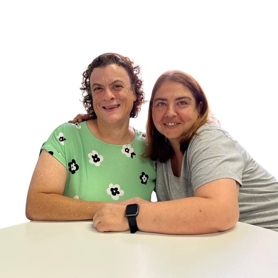 2 friends smiling and sitting close to each other, one has her arm around the other persons shoulders